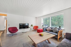 Un saló modern amb televisió plana, cadires vermelles i taronja, una taula de centre de fusta amb fruita i una finestra gran amb vistes a la vegetació.