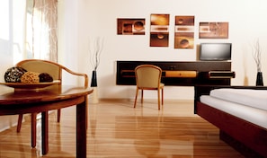 A hotel room with a wooden floor, a dining table with a bowl of fruit, a sofa, a TV, and artwork on the wall.