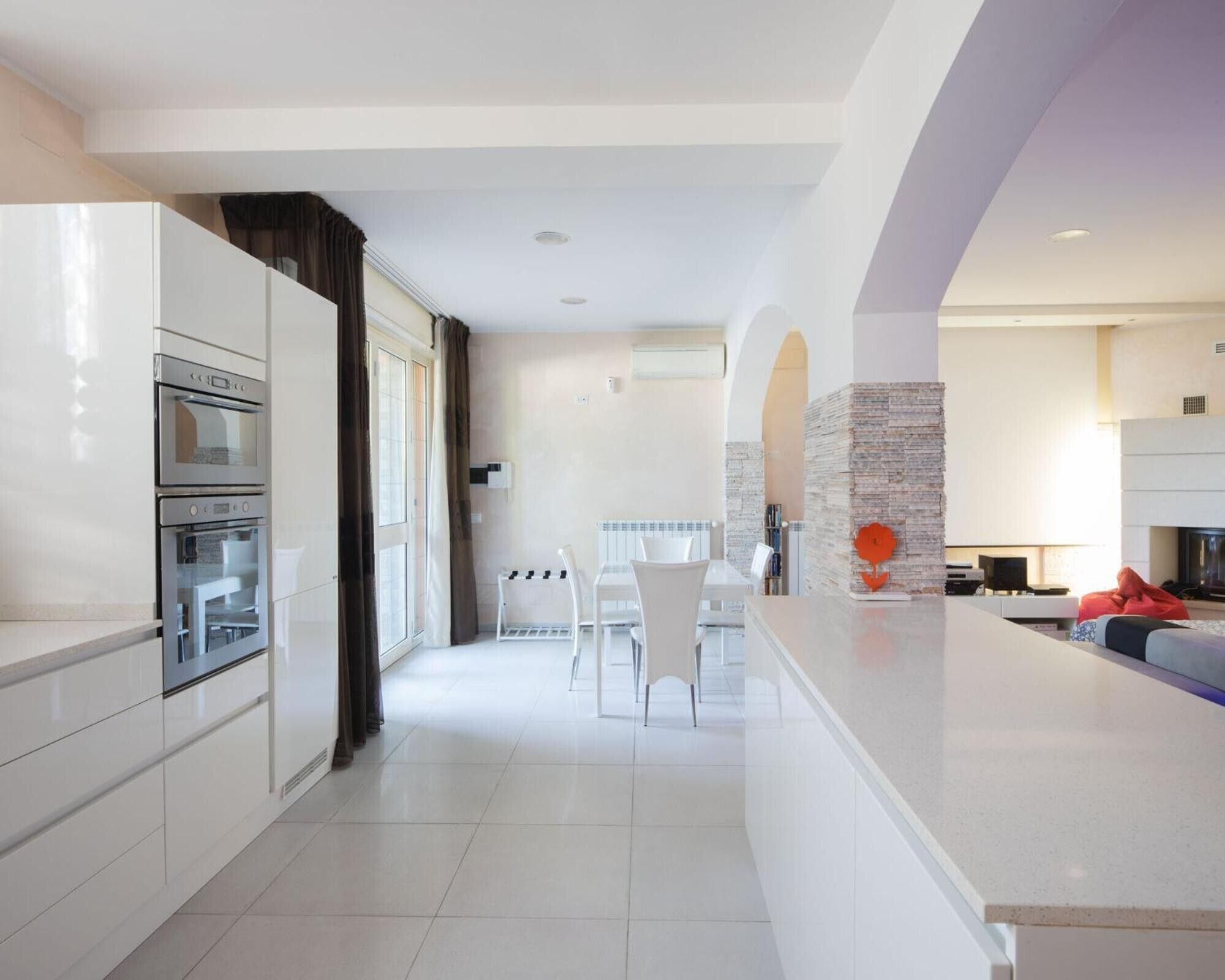 A modern kitchen with a built-in oven, a dining area with a white table and chairs, and a living area with a sofa and a fireplace.