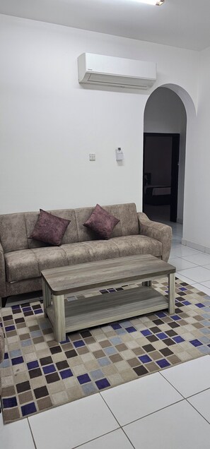 Una sala de estar con un sofá, dos cojines morados, una mesa de centro de madera, un tapete estampado y un equipo de aire acondicionado en la pared.