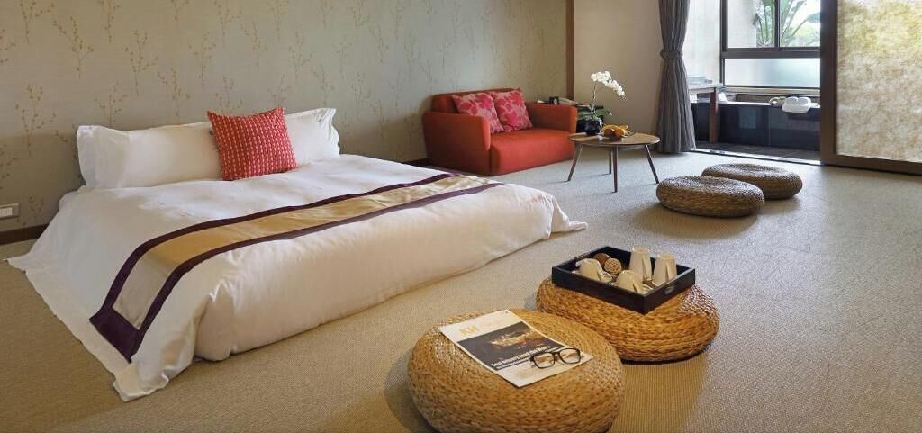 A hotel room with a large bed, a red sofa, a small table with a vase, and two round ottomans.