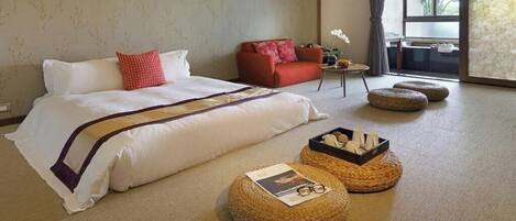 A hotel room with a large bed, a red sofa, a small table with a vase, and two round ottomans.