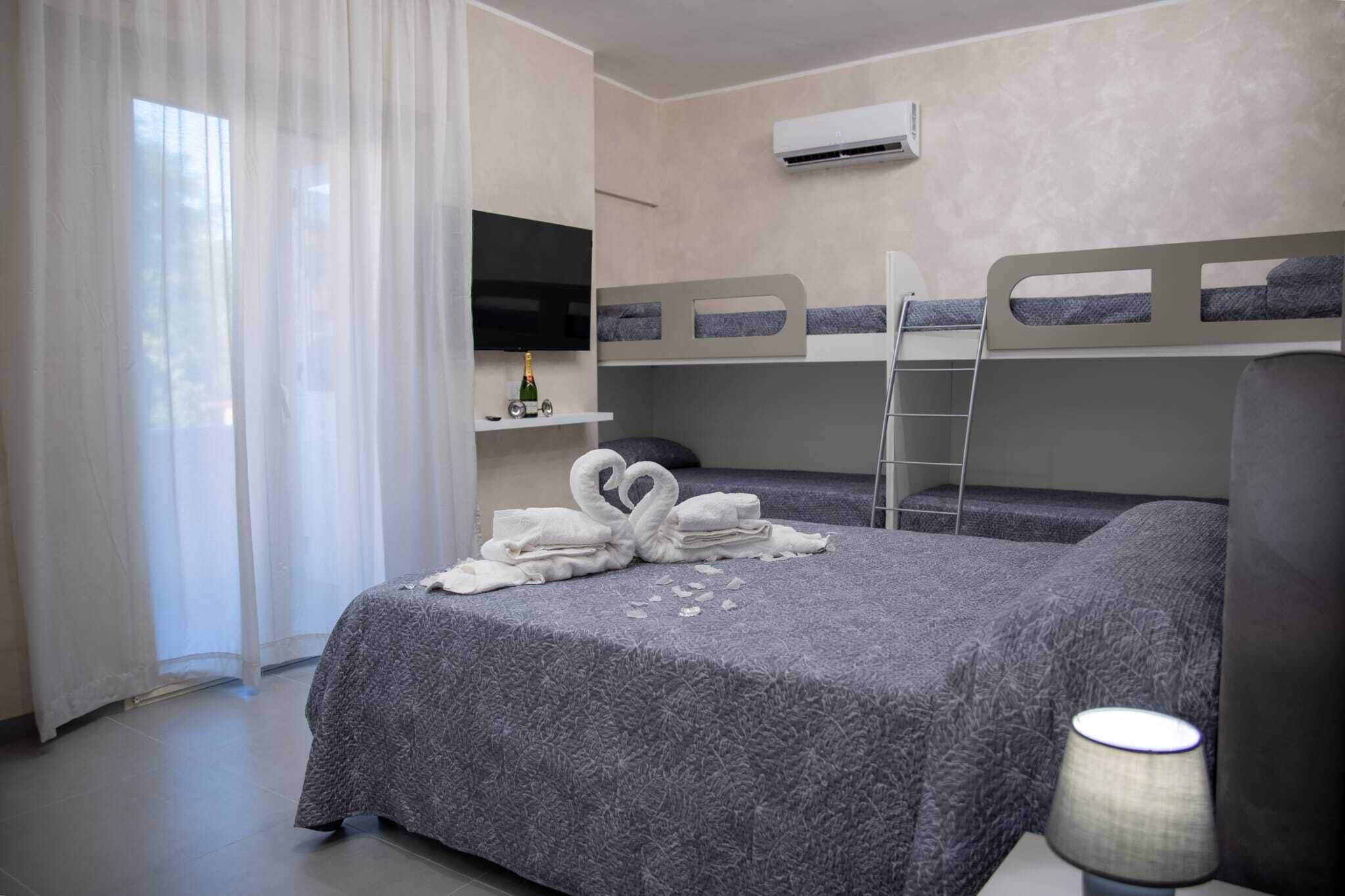 Ein Hotelzimmer mit Etagenbetten, einem Fernseher, einer Minibar und einem Fenster mit durchsichtigen Vorhängen.