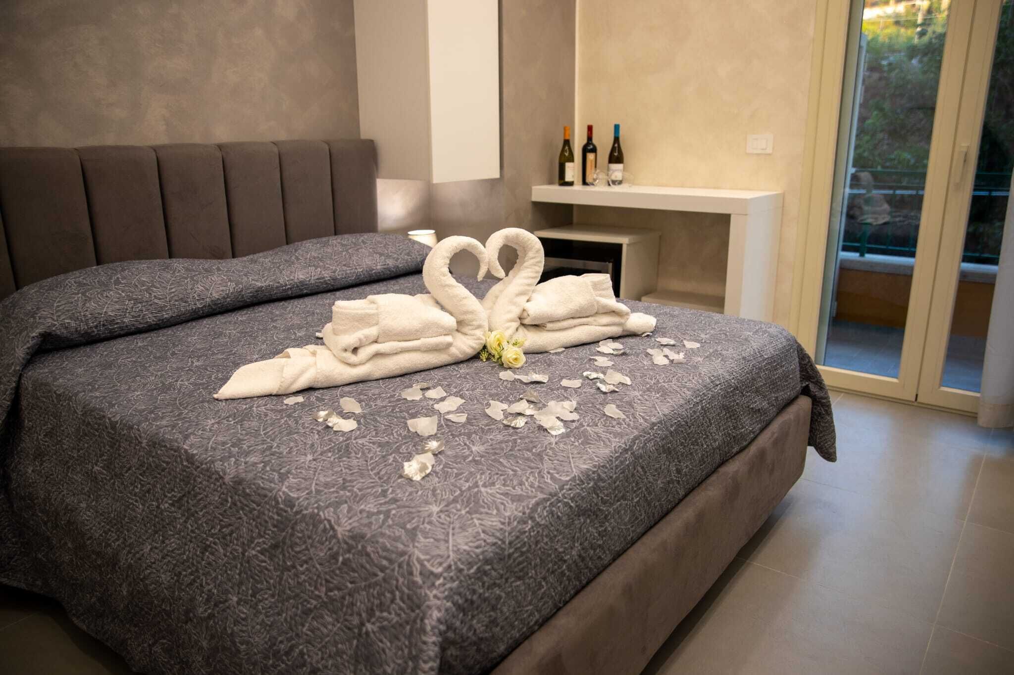 Uma cama com arranjo de toalhas em formato de coração, um pequeno vaso com uma flor e uma lareira com garrafas em uma prateleira.