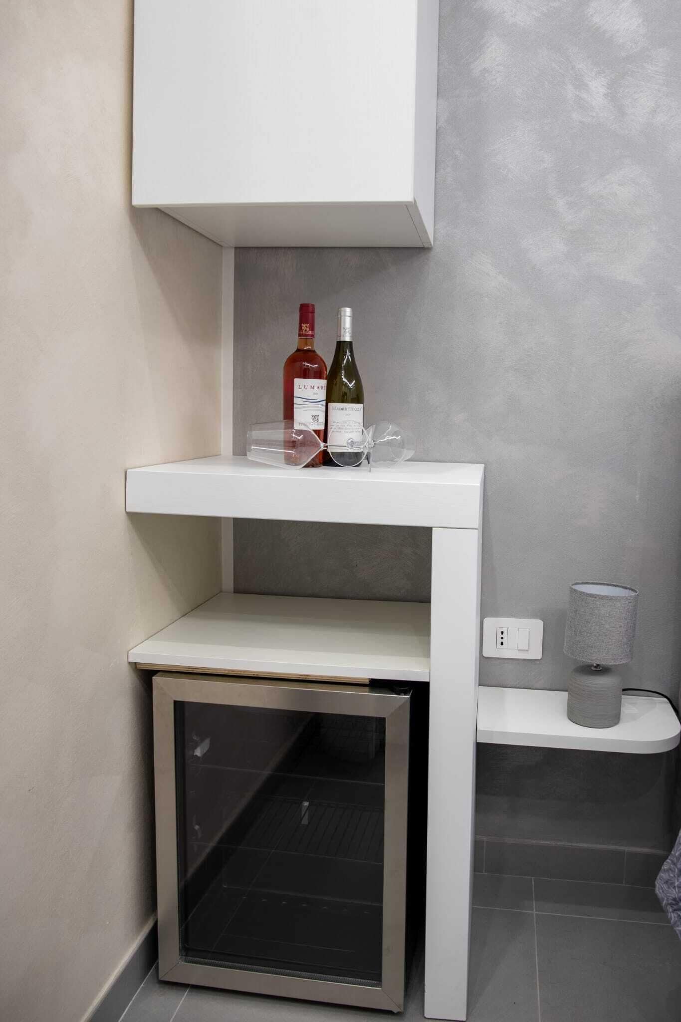 Un mueble blanco con una botella de vino y una copa encima, un estante pequeño debajo y un enchufe a la derecha.