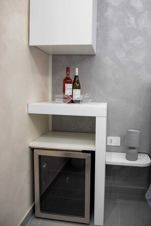 Un mueble blanco con una botella de vino y una copa encima, un estante pequeño debajo y un enchufe a la derecha.