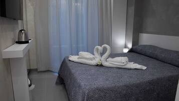 A neatly made bed with a towel arrangement, a bedside table with a kettle, and a window with sheer curtains.