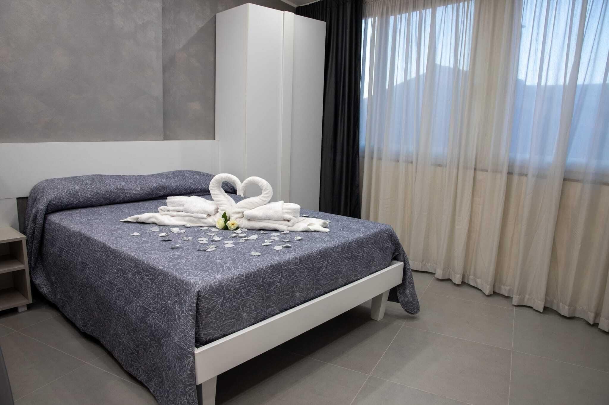 A neatly made bed with a heart-shaped towel arrangement, a white wardrobe, and a view of a building through sheer curtains.