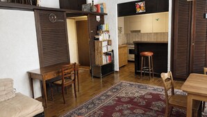 Ein Wohnzimmer mit Holzboden, einer Couch, einem Couchtisch, einem Teppich, einem Bücherregal und einer Küchenzeile mit Schränken und einer Arbeitsplatte.