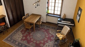 Ein Zimmer mit einem Holztisch, Stühlen, einem Teppich, einem Klavier, einem Schaukelstuhl und einem Fenster mit Jalousien.