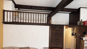 Ein gemütliches Zimmer mit einer Couch, einem Holztisch und einem Sessel. Ein Bücherregal mit Büchern und ein kleiner Tisch mit einer Lampe sind ebenfalls vorhanden.