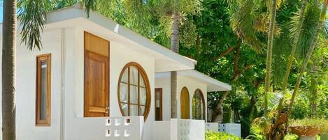 Un petit bâtiment d’un seul étage, à l’extérieur blanc, avec des portes et des fenêtres en bois, entouré d’une végétation luxuriante et de palmiers.