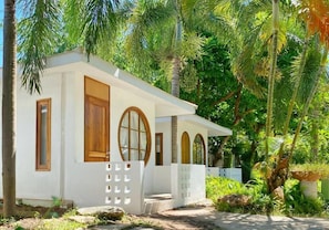 A small, single-story building with a white exterior, wooden doors, and windows, surrounded by lush greenery and palm trees. - Sangthong Resort (Chiang Klang)