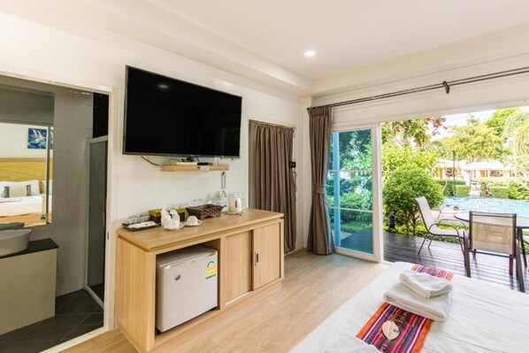 Habitación de hotel con cama, televisor, refrigerador pequeño, escritorio con computadora y vistas a una piscina y jardín.