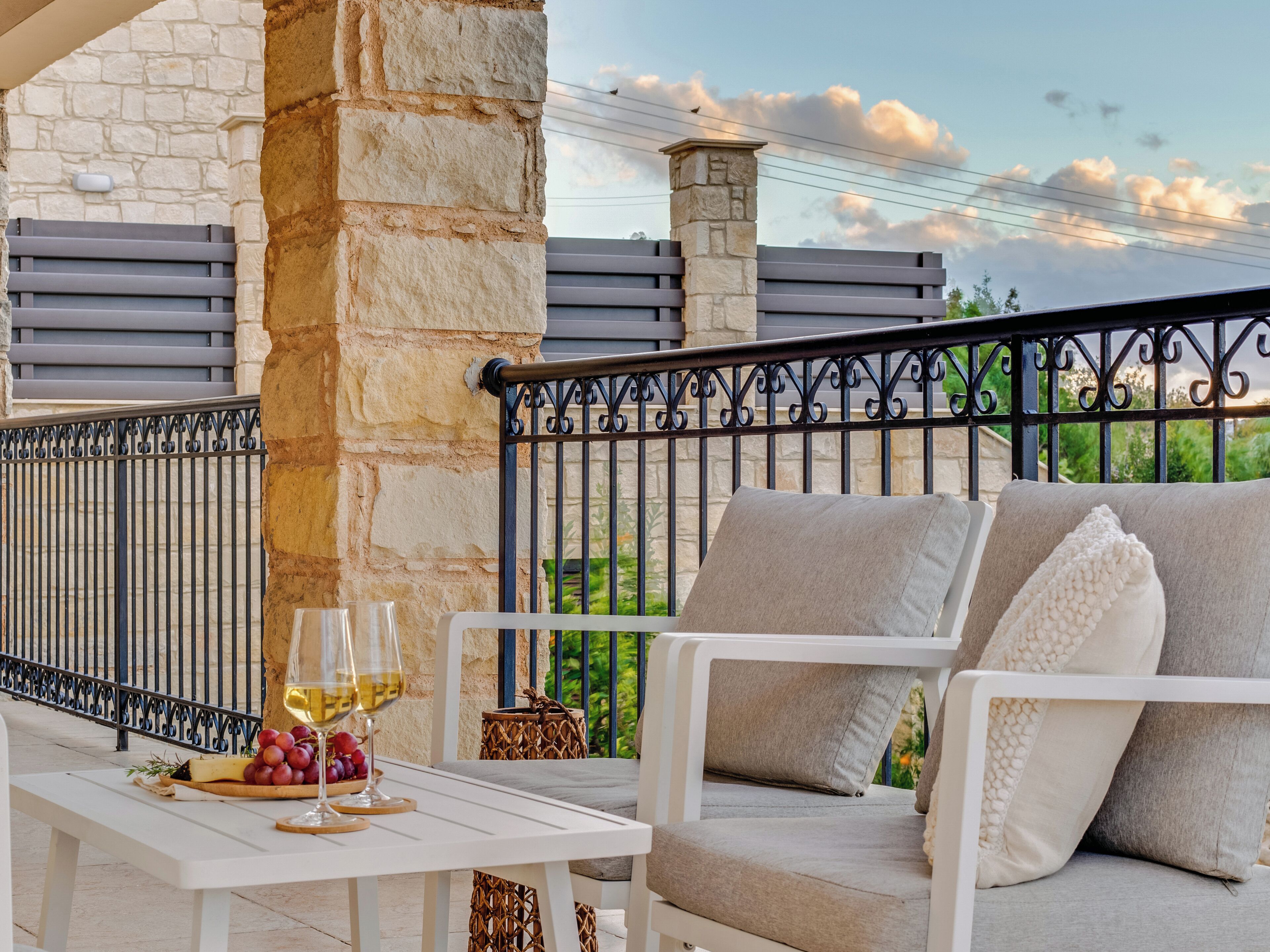 A balcony with a stone wall, a white table with a glass of wine, a wicker basket, and a white sofa with cushions.