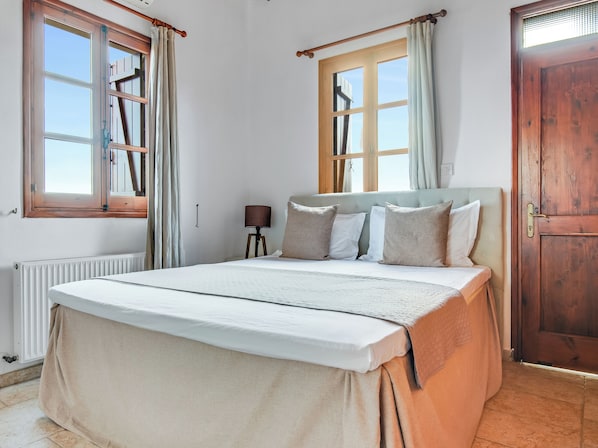 Un dormitorio con cama, mesita de noche, ventana con cortinas, puerta de madera y radiador.