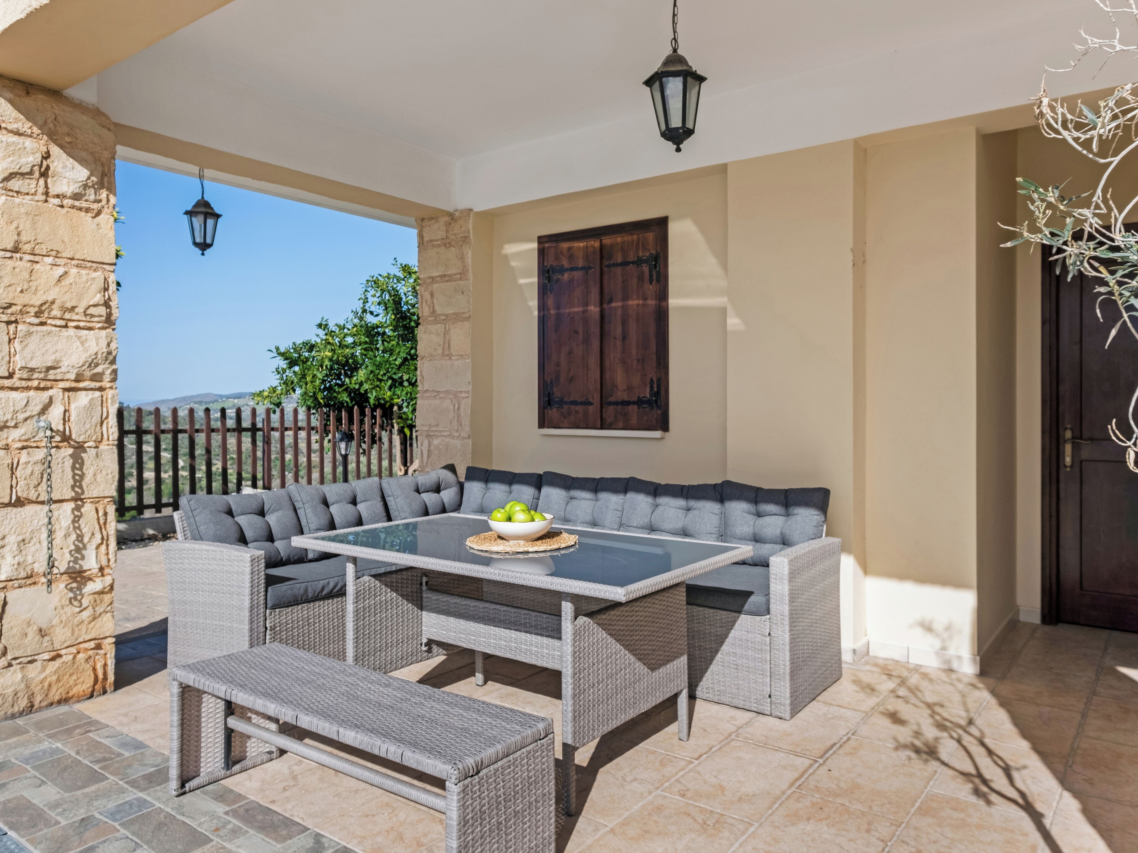 A covered outdoor seating area with a glass-top table, grey cushioned benches, and a wicker bench.