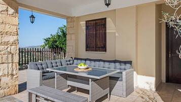 A covered outdoor seating area with a glass-top table, grey cushioned benches, and a wicker bench.
