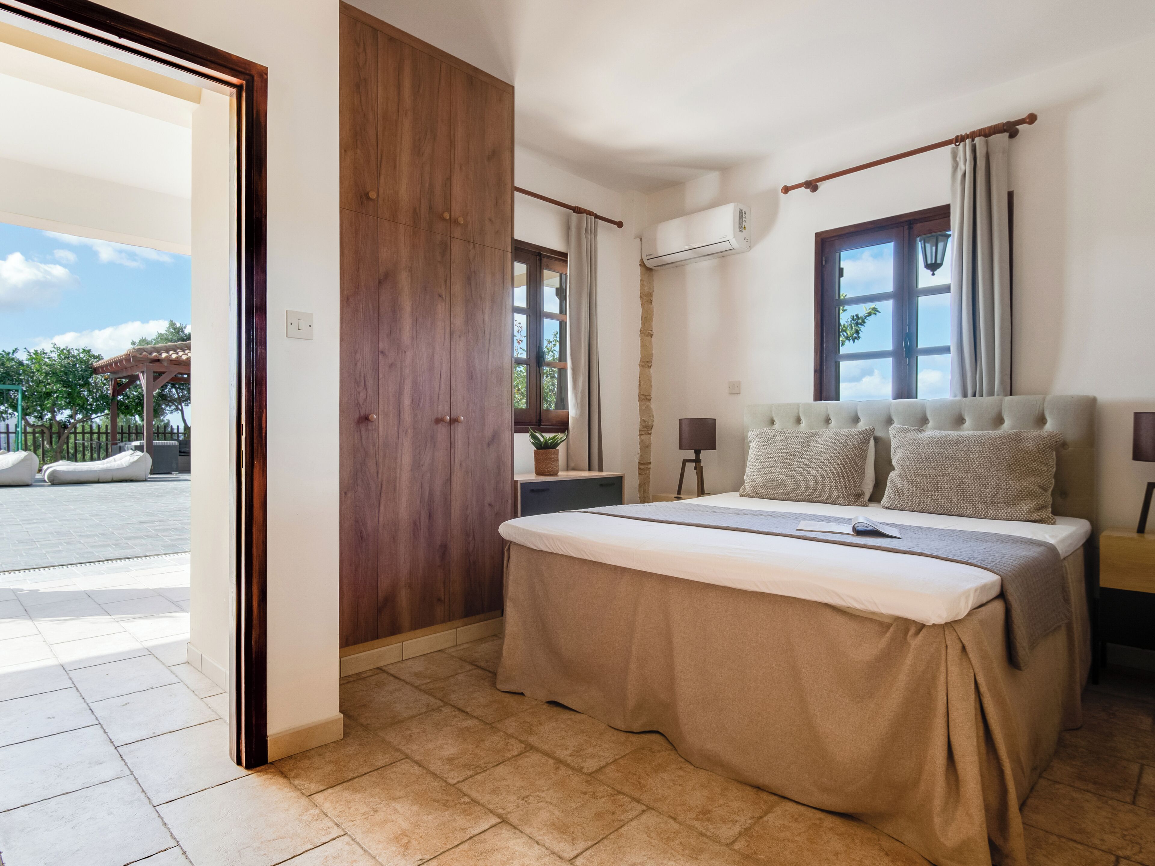 A bedroom with a bed, a wooden wardrobe, an air conditioning unit, a window with curtains, and a view of an outdoor area.