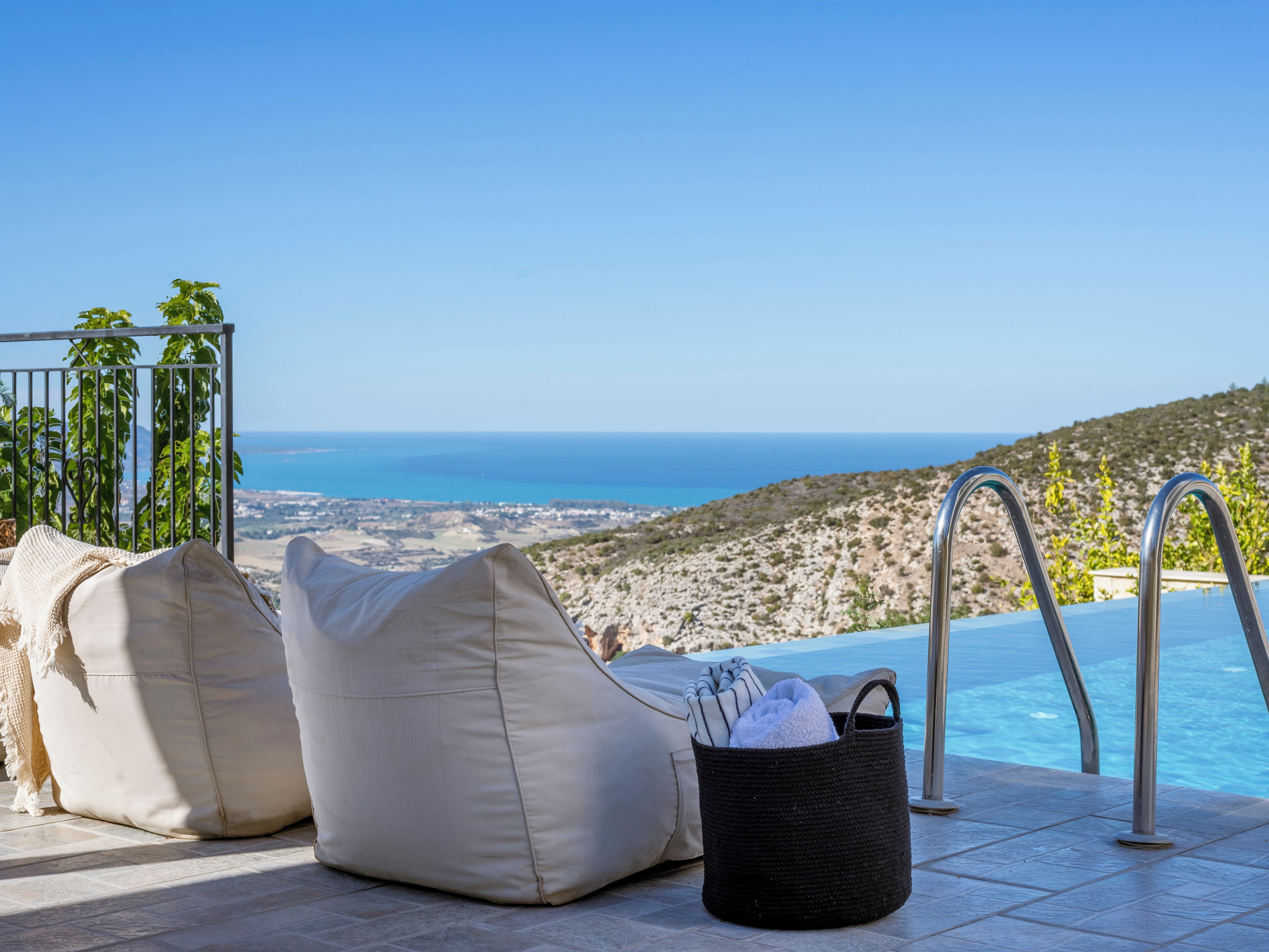 A poolside area with a view of the sea, two beige bean bags, and a black woven basket with towels.