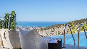 A poolside area with a view of the sea, two beige bean bags, and a black woven basket with towels.