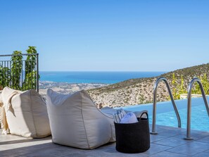 Zona junto a la piscina con vista al mar, dos puffs color beige y una cesta trenzada negra con toallas.