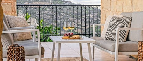 A balcony with two white chairs, a small table with a glass of wine, grapes, and a basket of bread, overlooking a hilly landscape.
