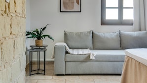 A room with a stone wall, a grey sofa, a small table with a plant, a framed picture, and a window with curtains. - Sea-view Retreat - Villas With Private Pools (Peristerona)