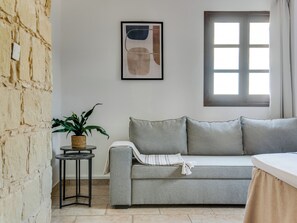 A room with a stone wall, a grey sofa, a small table with a plant, a framed picture, and a window with curtains.