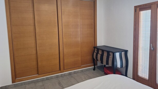 Un dormitorio con cama, armario, una mesita y una puerta con persianas.
