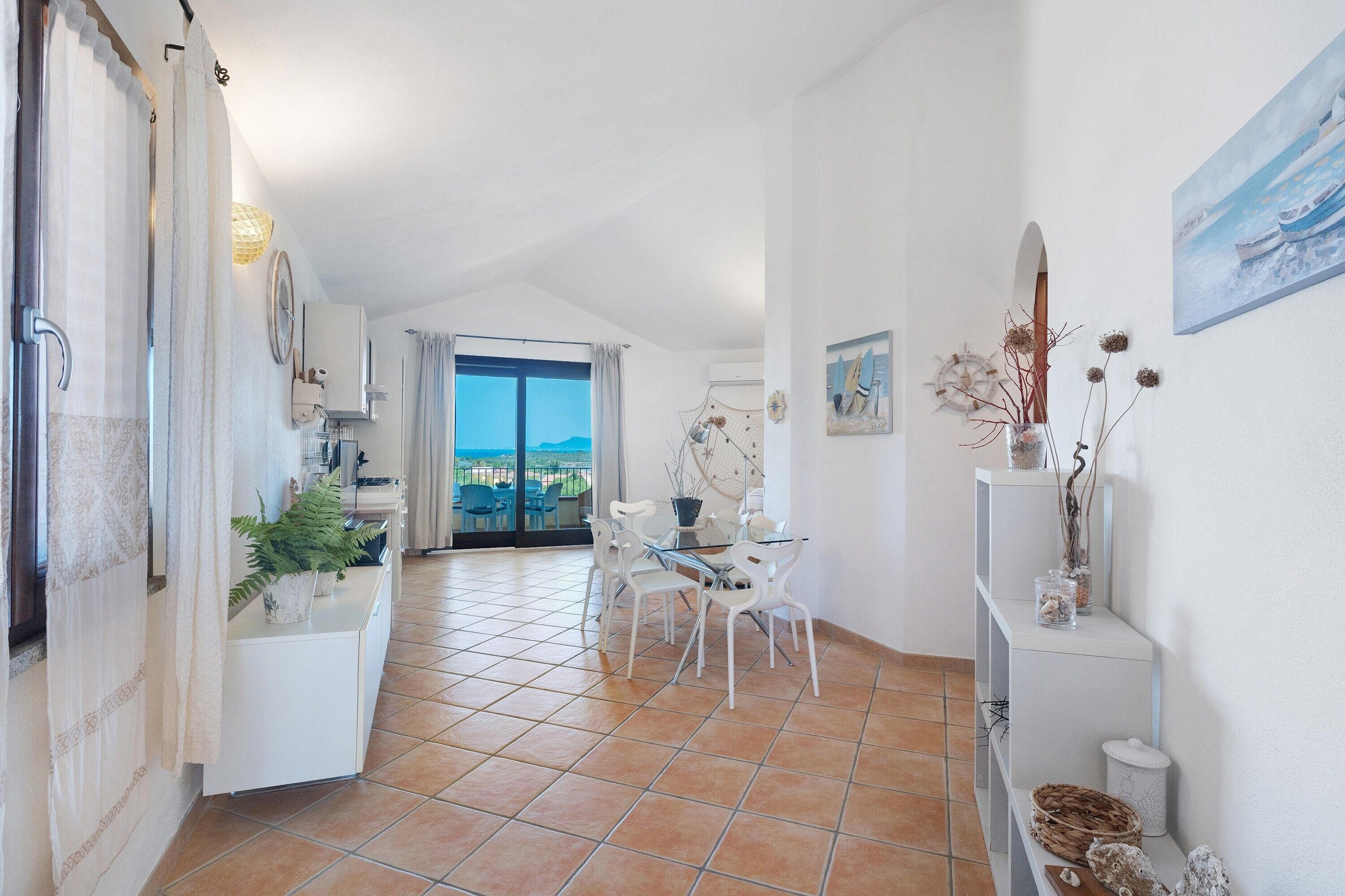 A spacious kitchen with a view to the outside, a dining area with white chairs, and a hallway leading to a balcony.