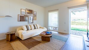 Ein Wohnzimmer mit einer Couch, einem Couchtisch und einem Teppich. An den Wänden hängen Bilder und durch das Fenster hat man einen Ausblick nach draußen.