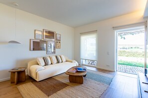 A living room with a sofa, coffee table, and a rug. There are framed pictures on the wall and a window with a view of the outside. - Tavira Sweet - Convento das Bernardas (Tavira)