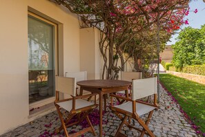 A patio with a round wooden table and four chairs, a glass door, and a garden with flowering plants. - Tavira Sweet - Convento das Bernardas (Tavira)