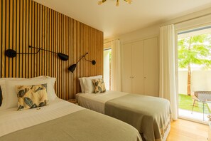 Ein Hotelzimmer mit zwei Betten, einer Wand aus Holzlatten, einer Deckenleuchte und Blick auf einen Garten durch eine Schiebetür.