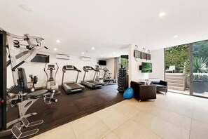 Academia com esteiras, elípticos e equipamentos de musculação. Há uma televisão na parede e um grande janelão com vista para o exterior.