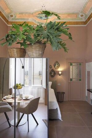 A room with a dining area, a bed, a wall-mounted light fixture, and a door with a decorative glass panel. - Charming Suite with Original Frescoes – Exclusive Suite Just 5 Minutes from Ortigia. (Siracusa)