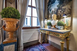 Ein Zimmer mit einem großen Fenster, einem dekorativen Tisch mit einer Büste und Topfpflanzen sowie einem Wandgemälde.
