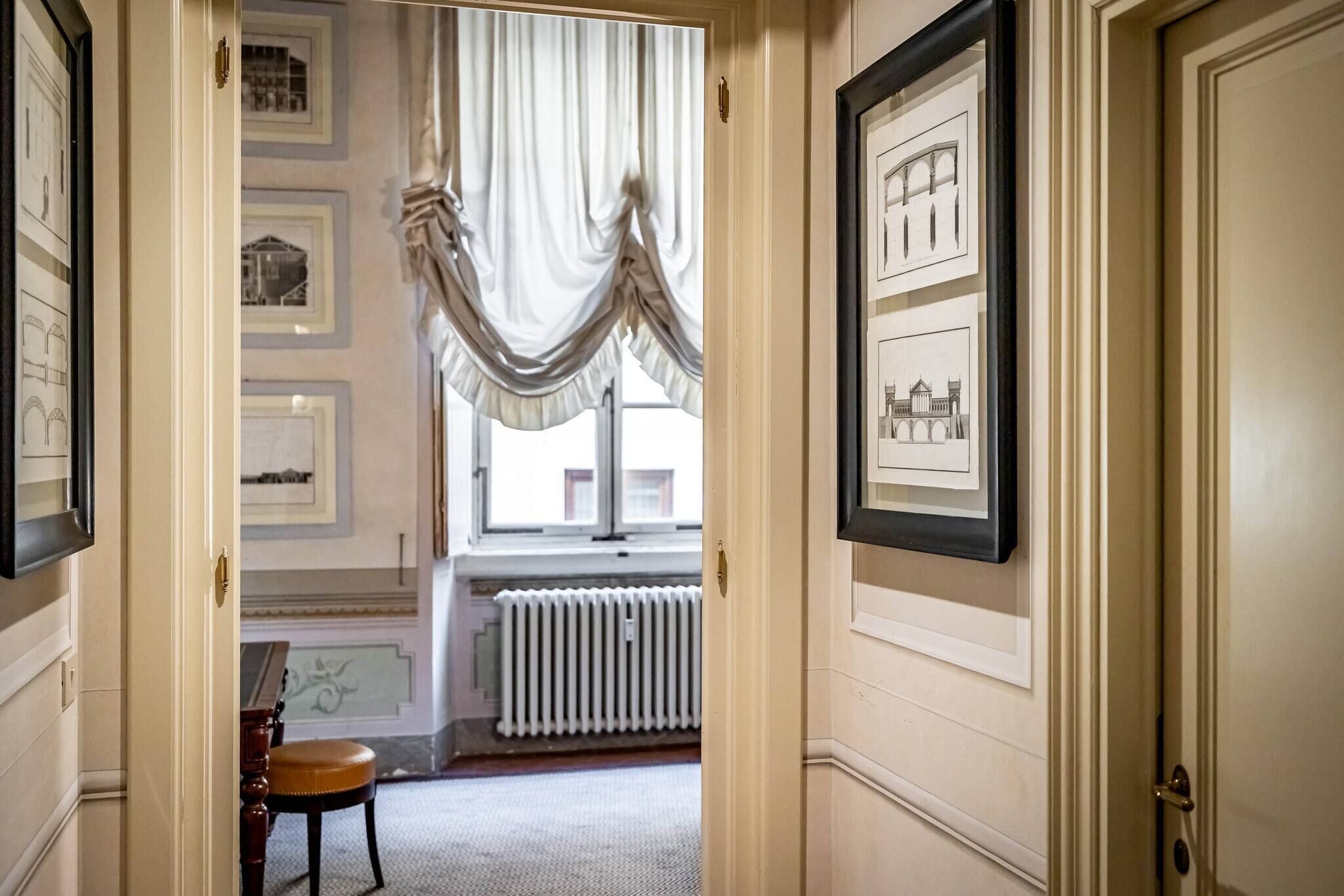 A hallway with framed architectural drawings on the walls, a radiator, and a window with draped curtains.