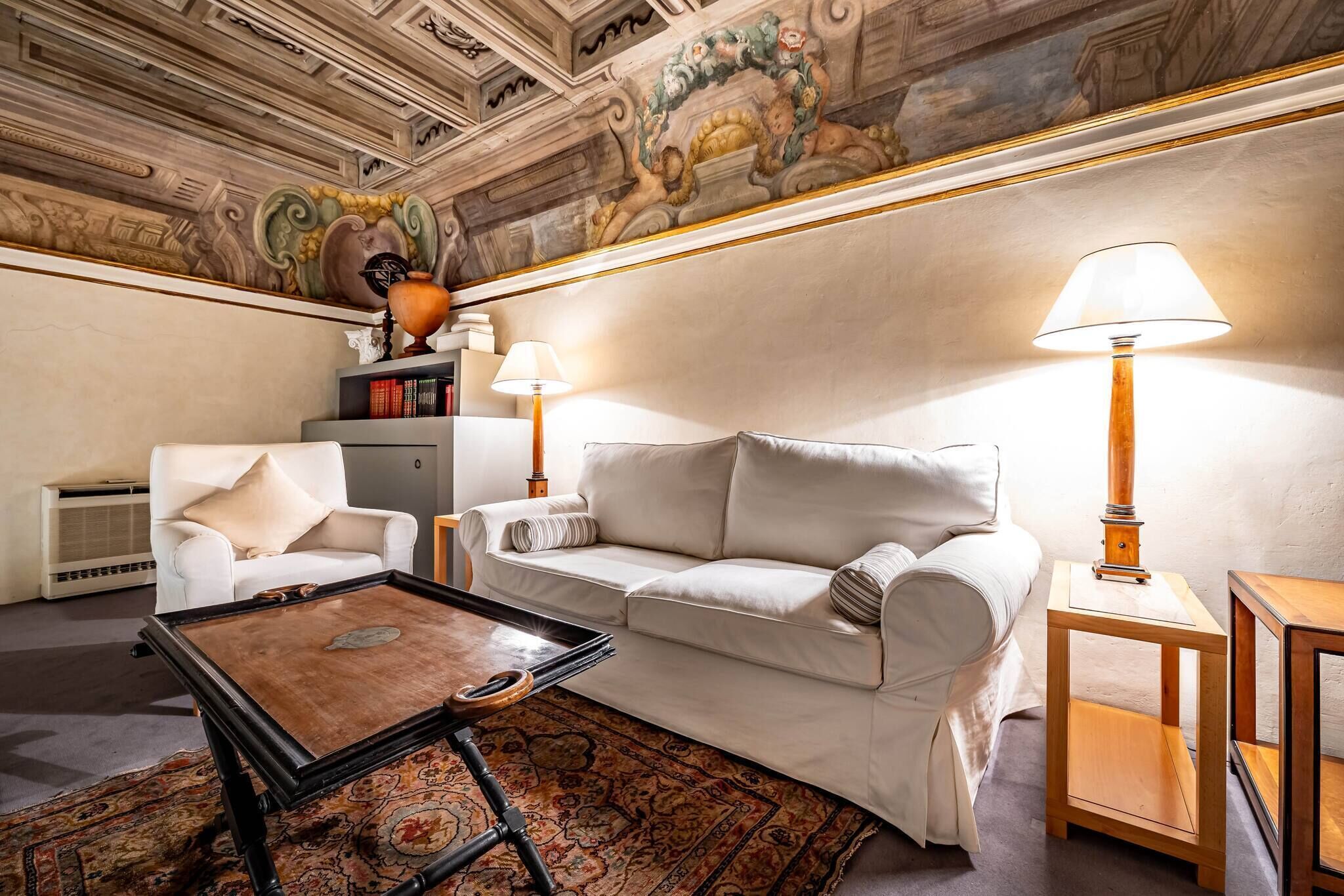 A living room with a white sofa, a wooden coffee table, and a lamp. There is a bookshelf and a decorative vase on a side table.