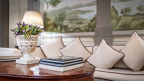 A round wooden table with a white decorative bowl containing purple flowers, a stack of books, and a white lamp. Behind the table, there is a beige sofa with white cushions and a large landscape painting on the wall. - Vigna Vecchia (Florence)