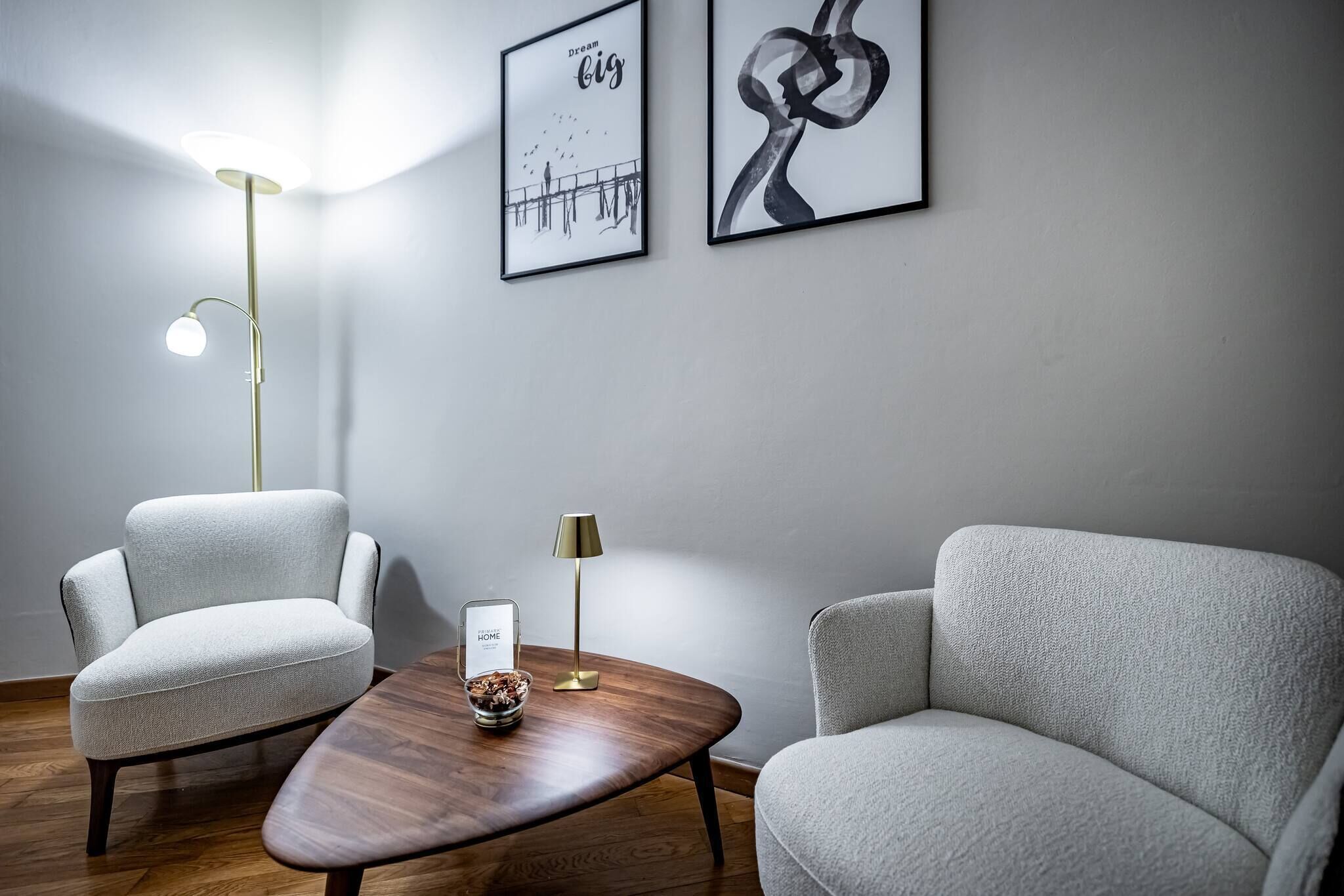 A modern room with two grey armchairs, a wooden coffee table, a floor lamp, and two framed artworks on the wall.
