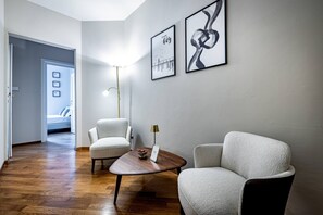 Ein modernes Wohnzimmer mit Holzboden, zwei weißen Sessel, einem kleinen runden Tisch mit einer Lampe und zwei gerahmten Kunstwerken an der Wand.