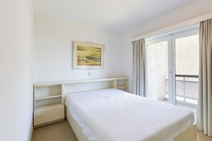 A bedroom with a bed, a bedside table, a shelf, a window with curtains, and a painting on the wall. - Ravelingen 0101+ G * (Koksijde)