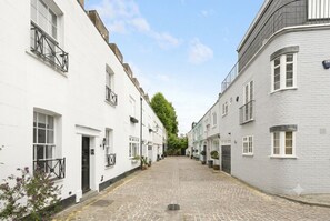 Eine enge Kopfsteinpflasterstraße, gesäumt von weißen Gebäuden mit schwarzen Geländern, strahlend blauer Himmel und ein Baum in der Ferne.