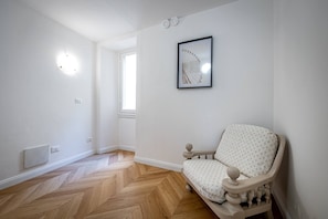 Una habitación con suelo de madera tipo espiga, un radiador blanco, una silla blanca con cojín estampado y un cuadro en la pared.