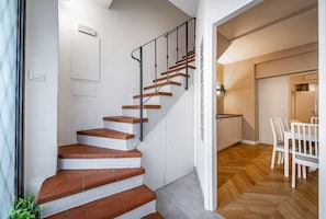 Una escalera con paredes blancas y escalones de madera que conduce a una zona de cocina con una mesa y sillas.