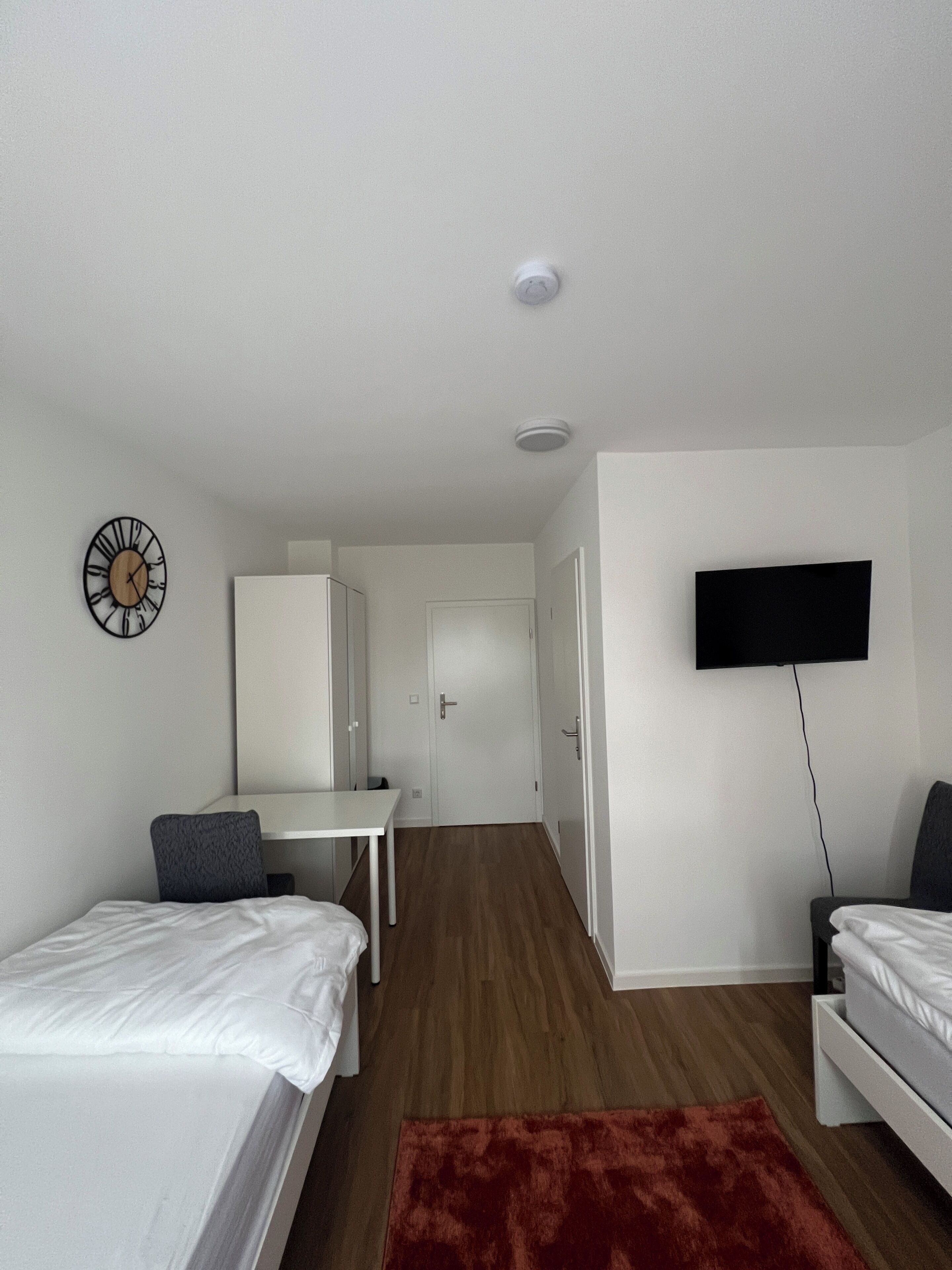 A hotel room with two beds, a desk, a TV, a clock, and a wardrobe.