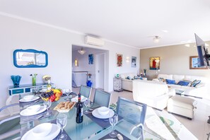 Ein modernes Wohnzimmer mit einem Esstisch aus Glas, gedecktem Tisch für eine Mahlzeit, einem weißen Sofa und einem flachbildfernseher an der Wand.
