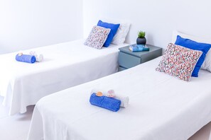 Zwei Einzelbetten mit weißen Bettwäsche und blauen Kissen, ein kleiner Nachttisch mit einem Buch und einer Topfpflanze, und eine weiße Wand im Hintergrund.